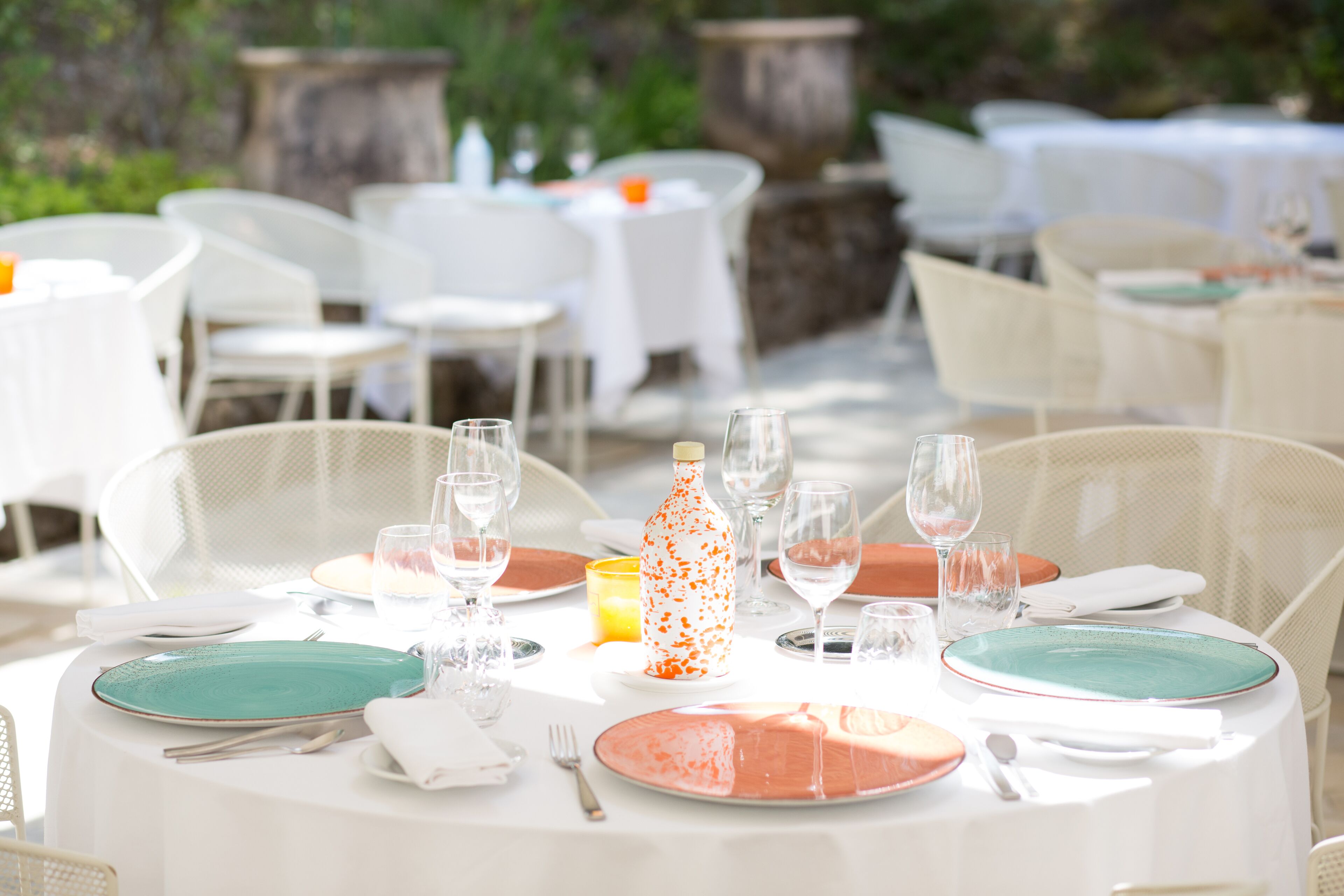 A round outdoor dining table set with white tablecloths, green and orange plates, and clear glassware.