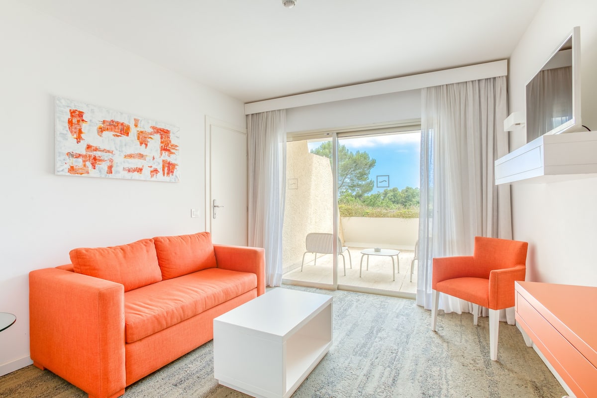 Une chambre d’hôtel moderne dotée d’une grande porte-fenêtre coulissante donnant sur un balcon, d’un canapé orange vif, d’une table basse blanche et d’un fauteuil assorti.