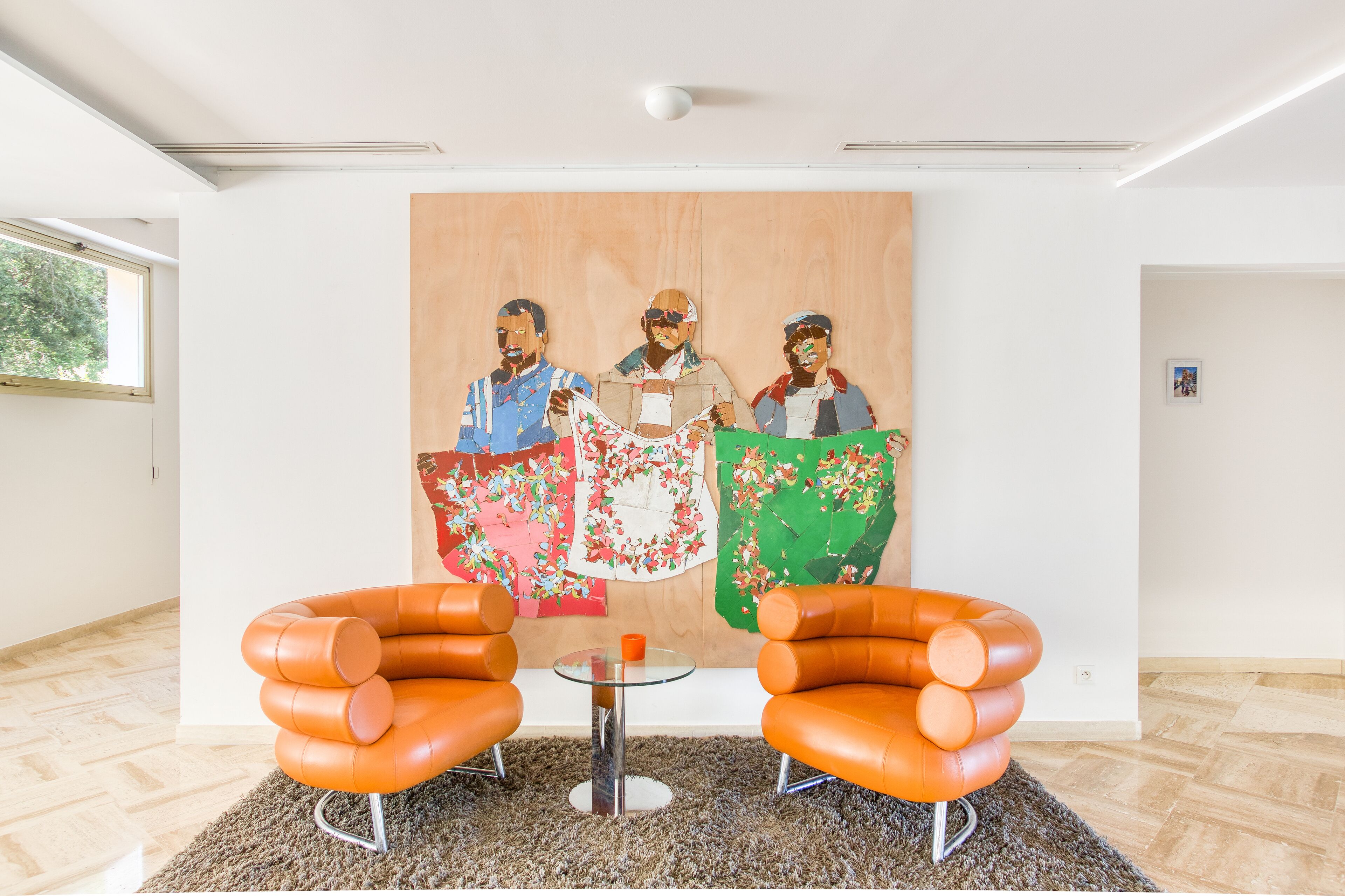 A room with a large painting, two orange chairs, and a small table.
