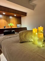 Un dormitorio con una cama, cabecera de madera, mesita de noche con una planta y tres velas encendidas.