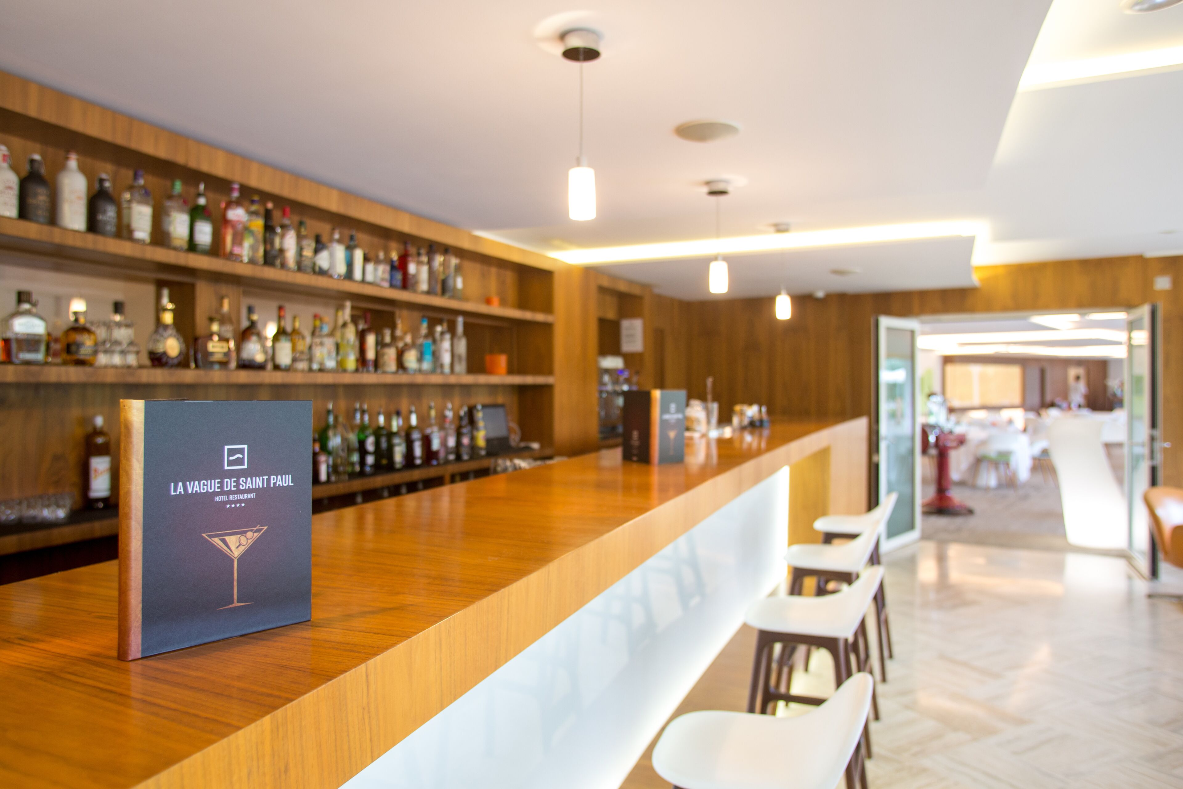 A bar area with a wooden counter, bar stools, and shelves of liquor bottles. There is a sign with the text "LA VAGUE DE SAINT PAUL HOTEL RESTAURANT."