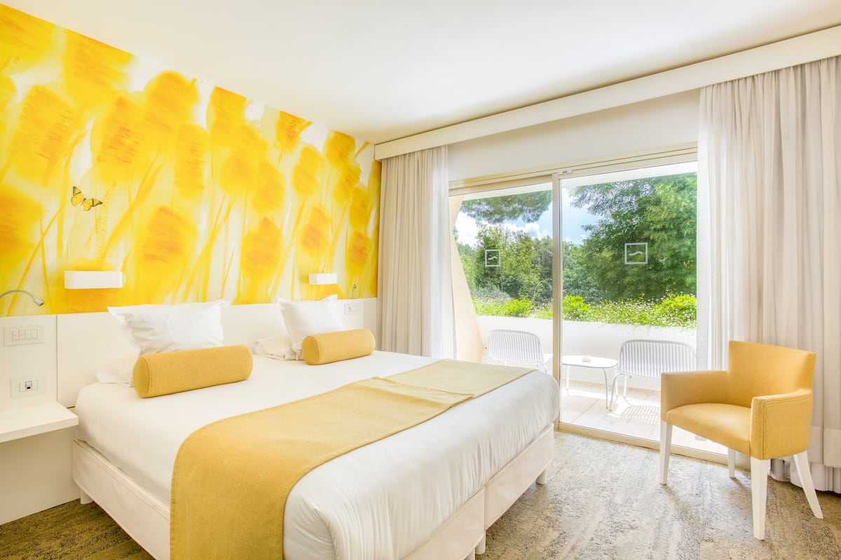 Une chambre d’hôtel avec un grand lit, un mur d’accent jaune, un balcon avec une chaise et une vue sur la verdure.