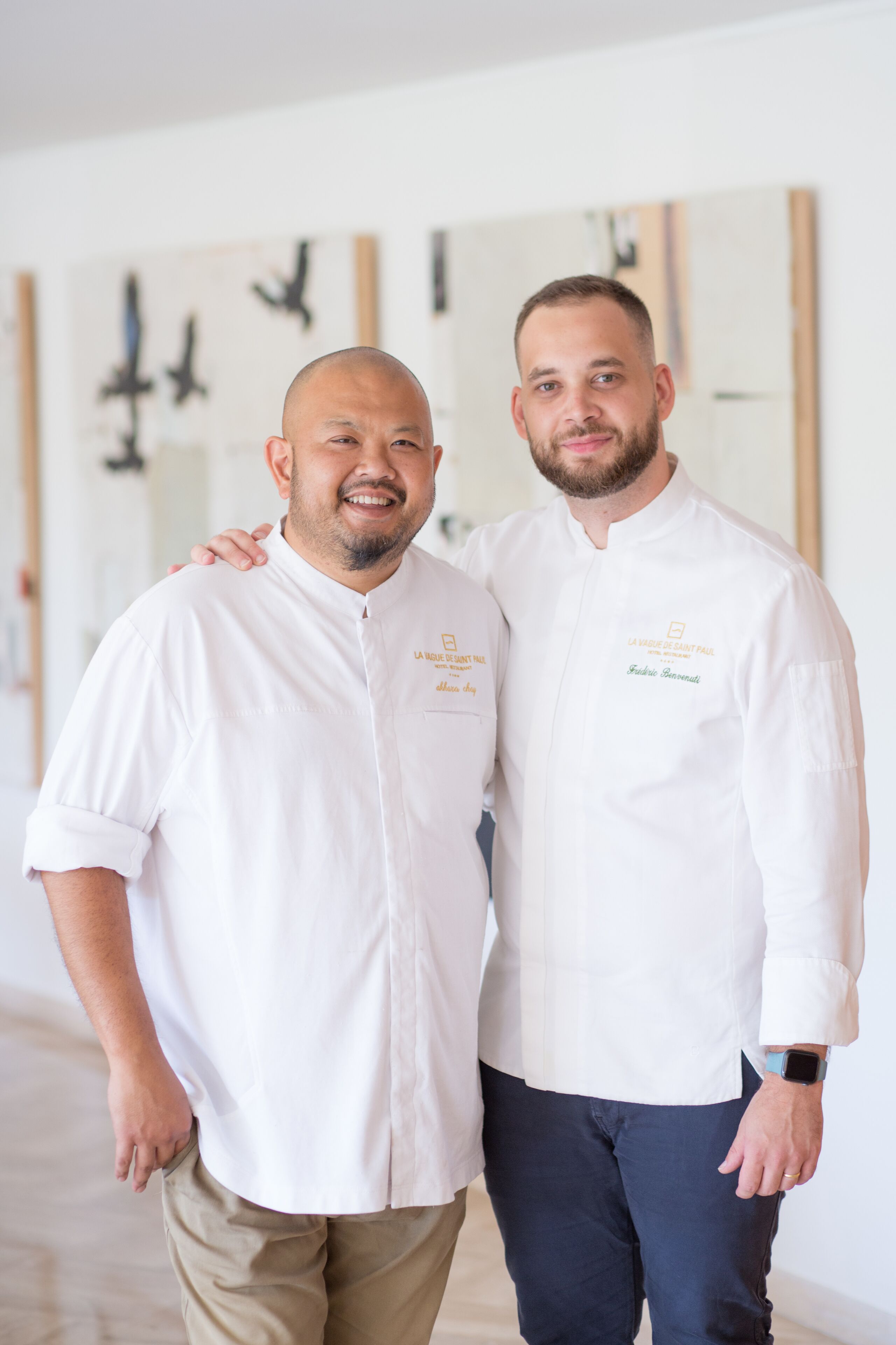 Dos hombres con uniformes de chef de pie en un pasillo con un muro espejado y obras de arte.