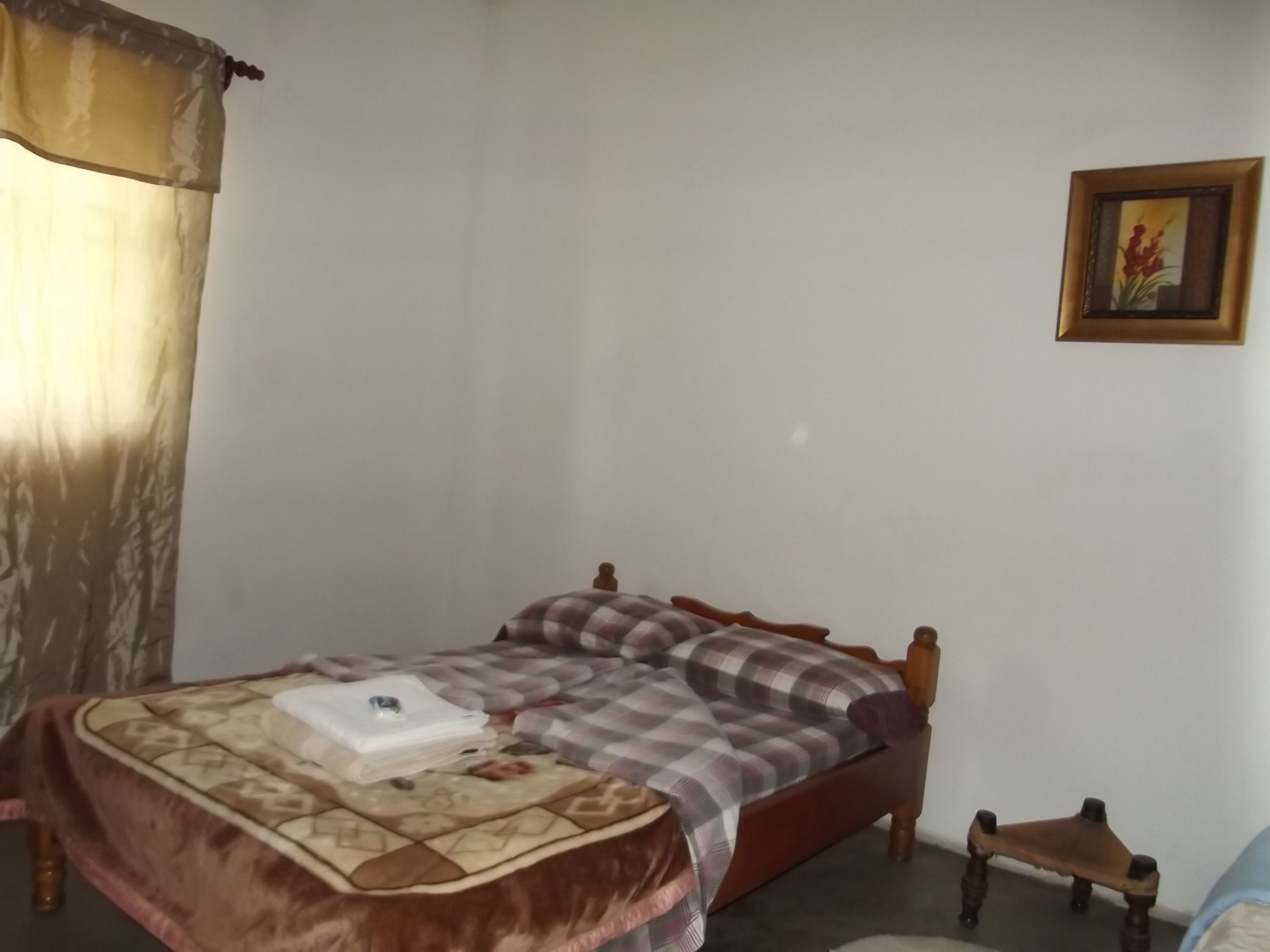 Een slaapkamer met een bed, nachtkastje, stoel, gordijn en een aan de muur hangend schilderij.