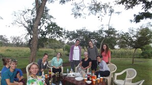 A group of people seated around a table with food and drinks, outdoors with trees in the background.