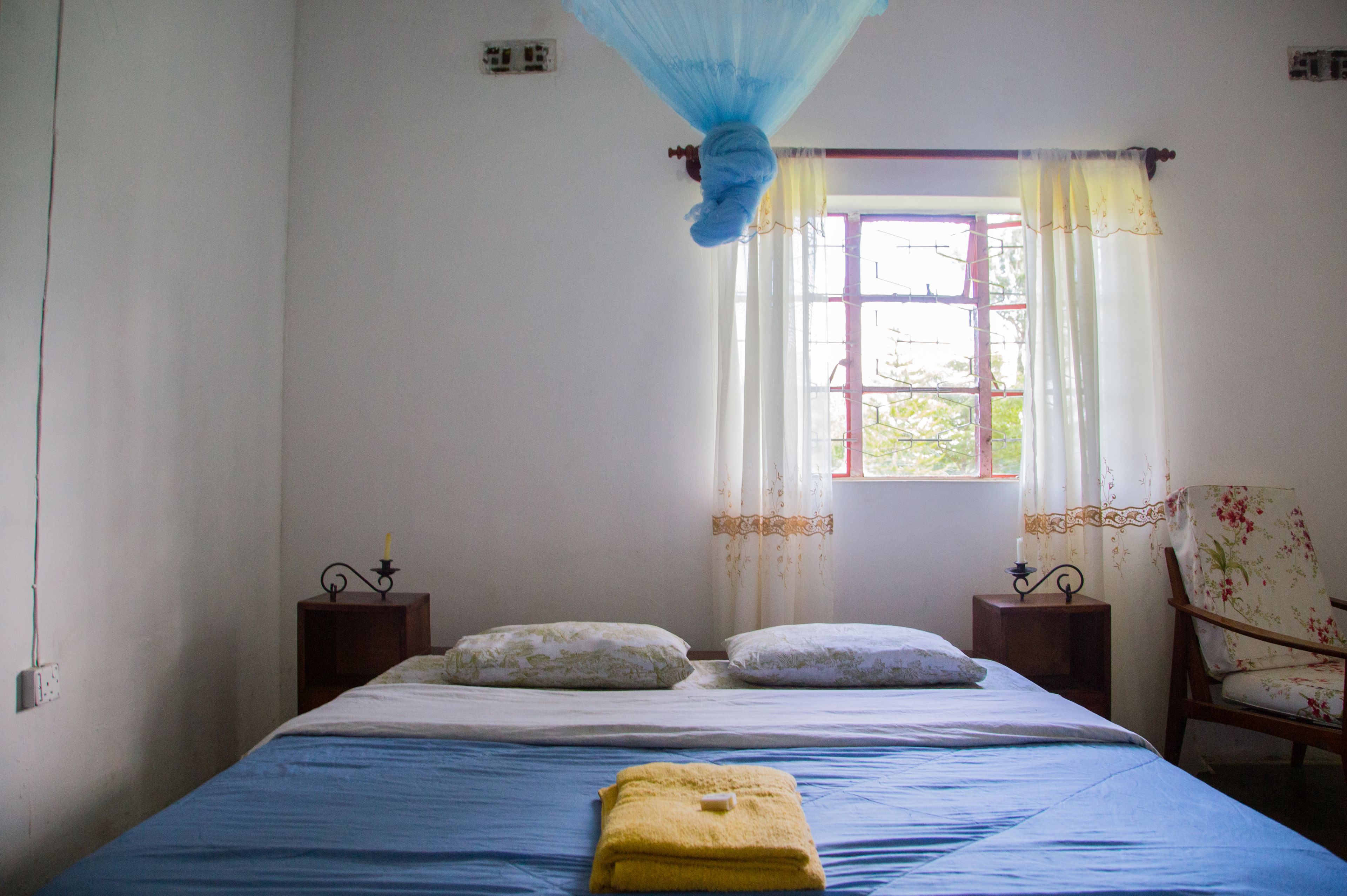 Un dormitorio con cama, dos mesitas de noche, una silla, una ventana con cortinas y una mosquitera.