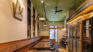 A restaurant interior with wooden benches, pendant lighting, a ceiling fan, and a counter with a beverage sign. - APA Hotel Ginza Shintomicho Ekimae (Tokyo)