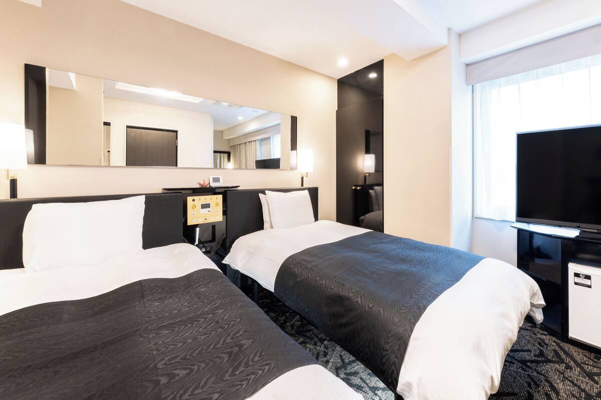 A hotel room with two beds, a TV, a desk with a lamp, a mirror, and a window.