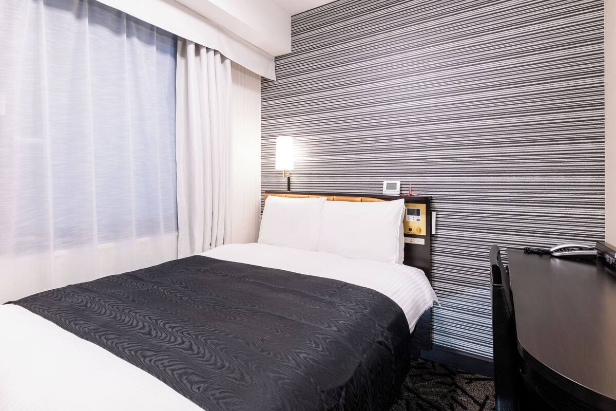 A hotel room with a bed, a desk, a lamp, and a wall with a striped pattern.