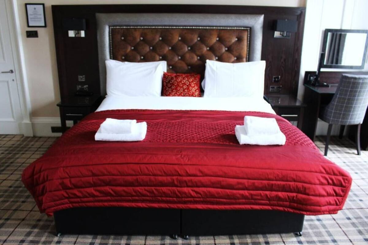 Habitación de hotel con una cama grande, un cabecero oscuro, una colcha roja, almohadas blancas y una almohada decorativa roja. Hay un escritorio con una silla y un espejo en la esquina.