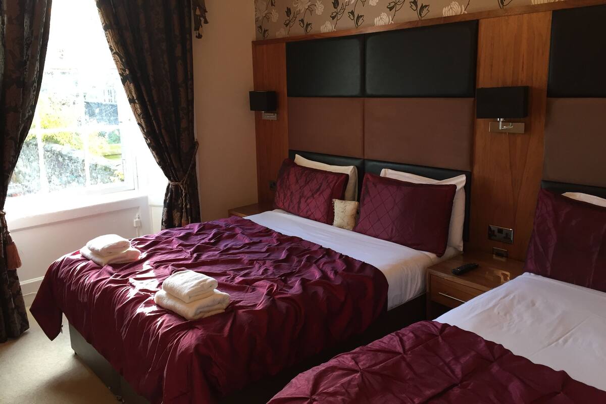 Habitación de hotel con dos camas, un ventanal con cortinas, papel tapiz con motivos florales y cabecero de madera.