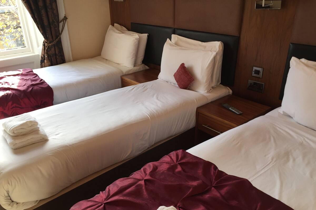 Habitación de hotel con dos camas individuales, cada una con sábanas blancas y una manta roja. Se observa un cabecero de madera, una mesita de noche con un control remoto y una ventana con cortinas.
