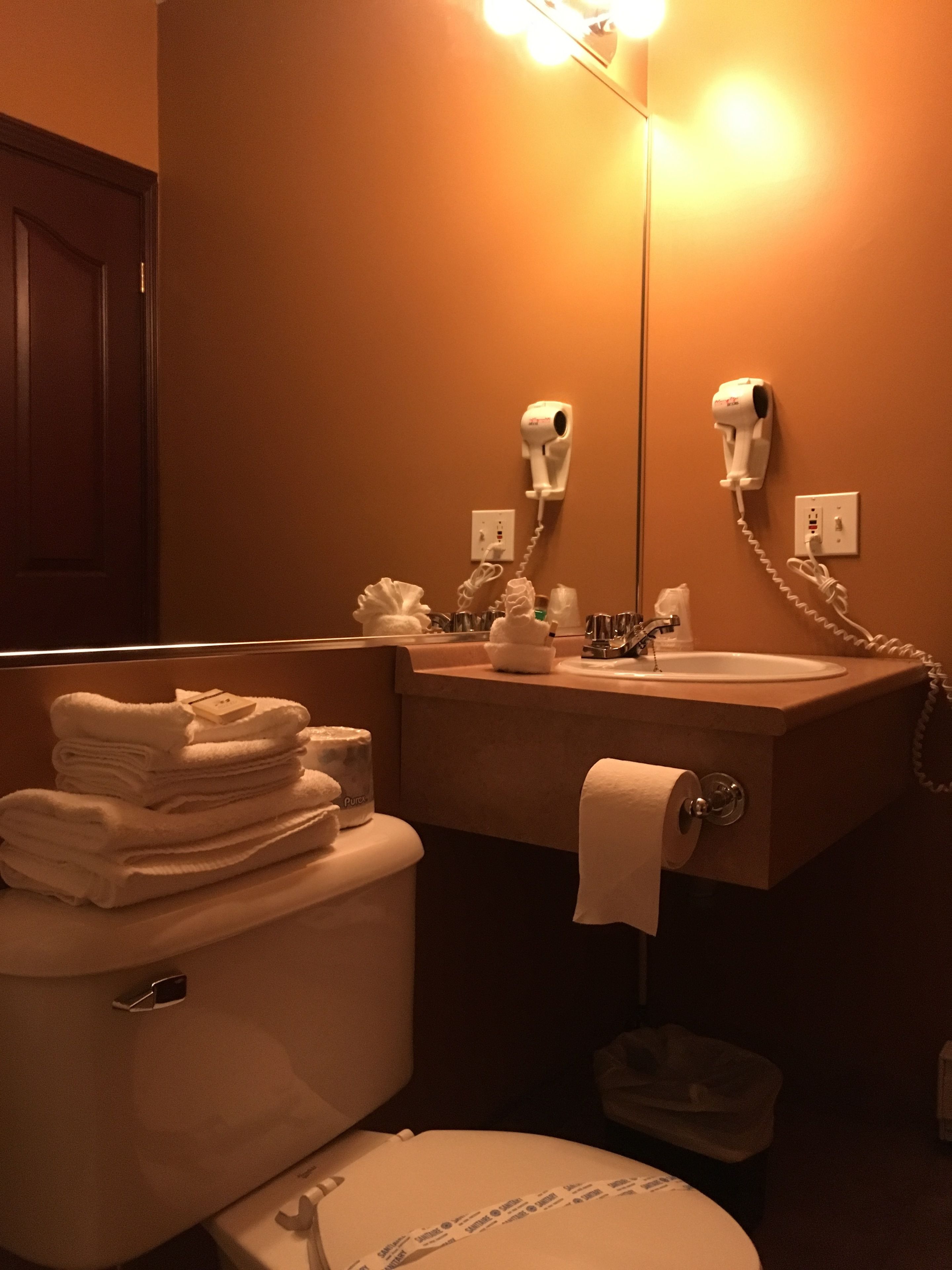 A bathroom with a toilet, sink, and hairdryer mounted on the wall. There are towels stacked on the toilet and a trash bin.