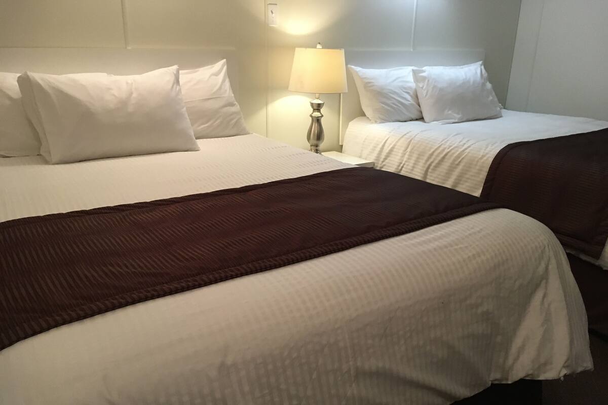 Habitación de hotel con dos camas, cada una con ropa de cama y almohadas blancas. Una manta marrón está sobre una de las camas. Se ve una mesita de noche con una lámpara en el lado izquierdo de la habitación.