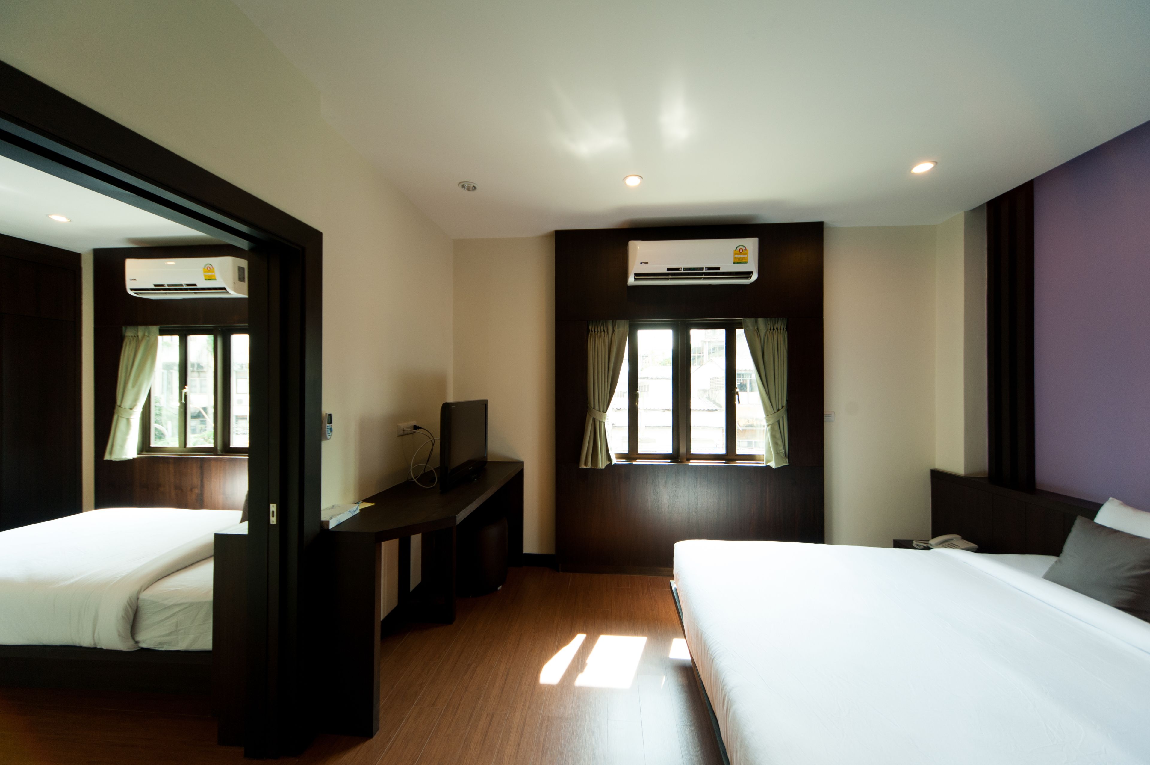 A hotel room with two beds, a desk with a TV, an air conditioning unit, and a window with curtains.