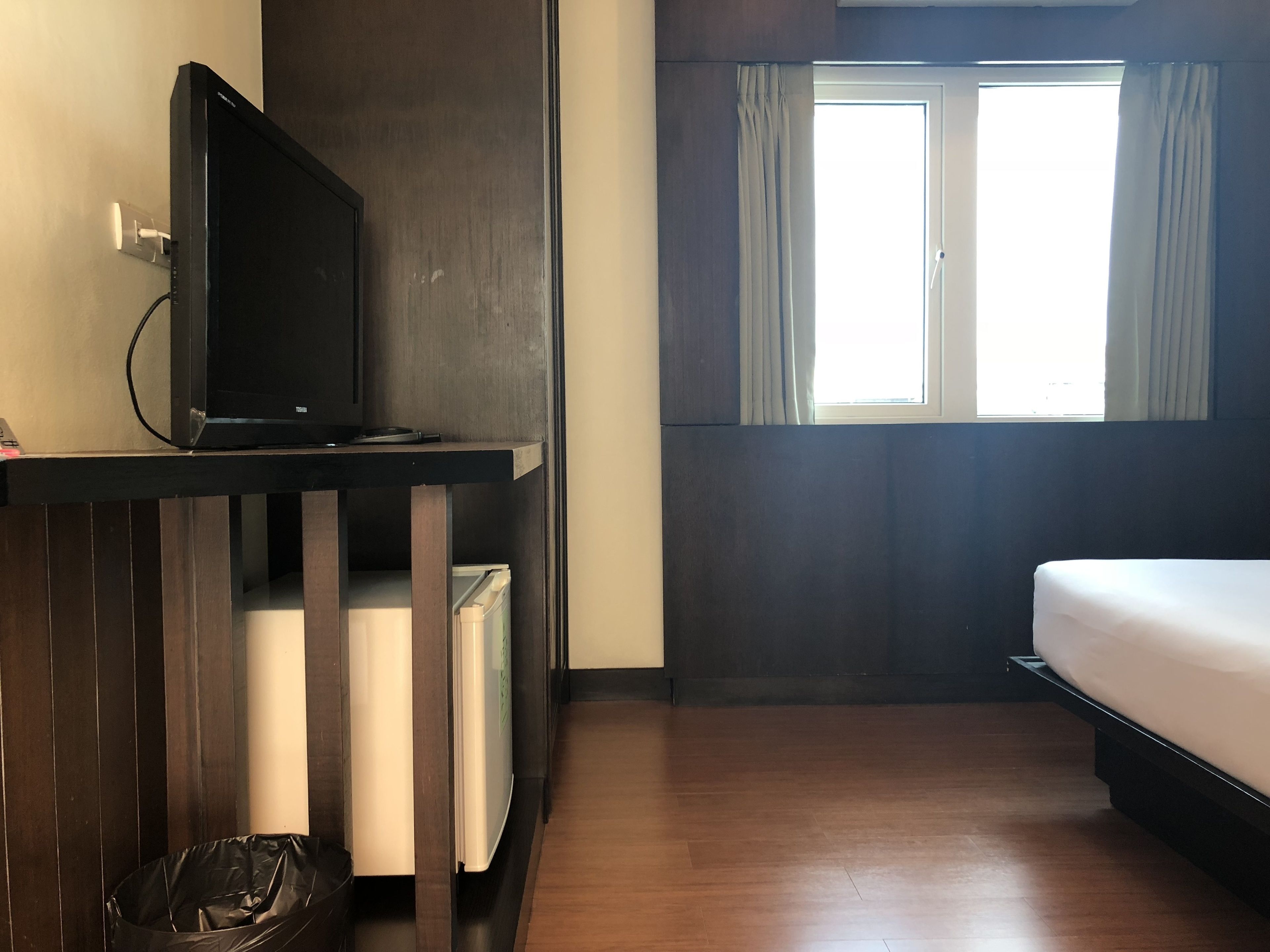 Ein Hotelzimmer mit Bett, Fernseher auf Holzständer, Fenster mit Vorhängen und Mülleimer.