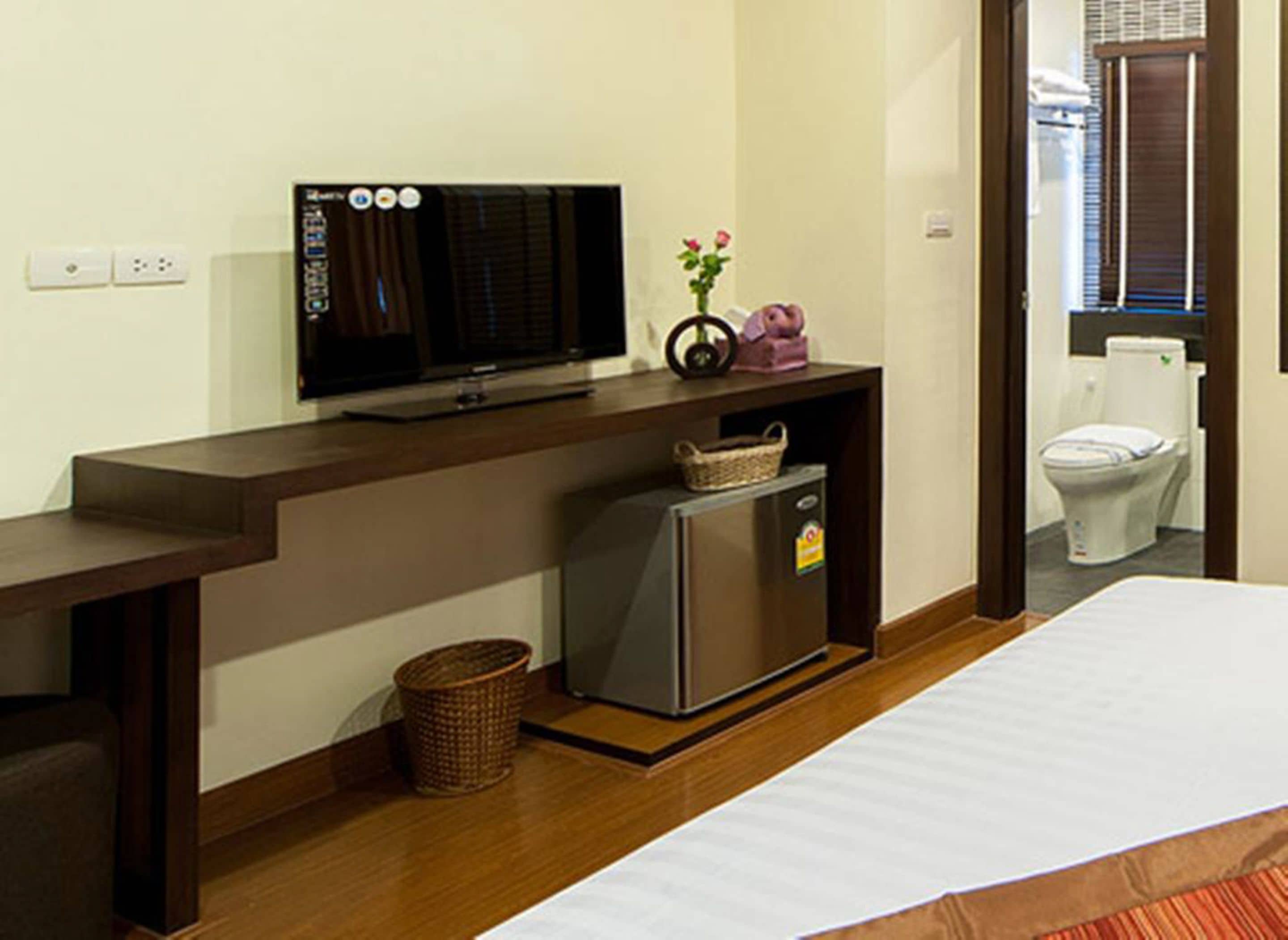 A hotel room with a wooden desk, a television, a small refrigerator, a basket, and a toilet visible through an open door.
