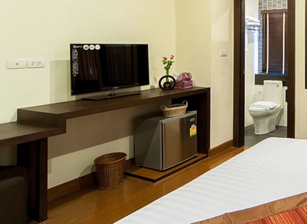 Habitación de hotel con escritorio de madera, televisor, refrigerador pequeño, cesta y un inodoro visible a través de una puerta abierta.