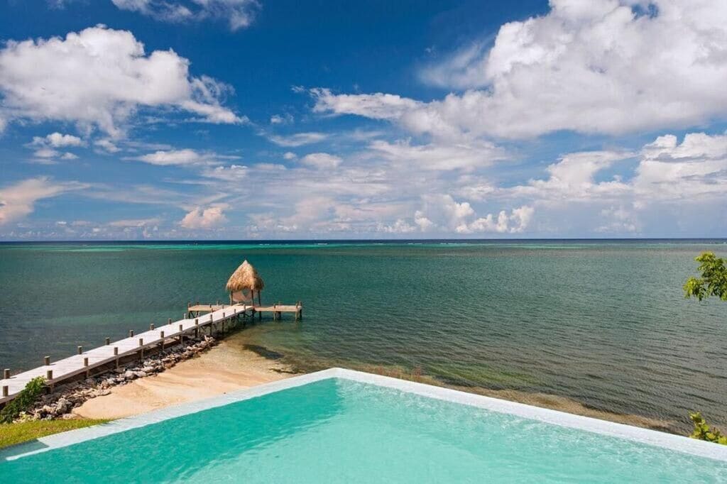 A clear blue sky, a wooden deck leading to a thatched-roof structure, and a pool with clear water.