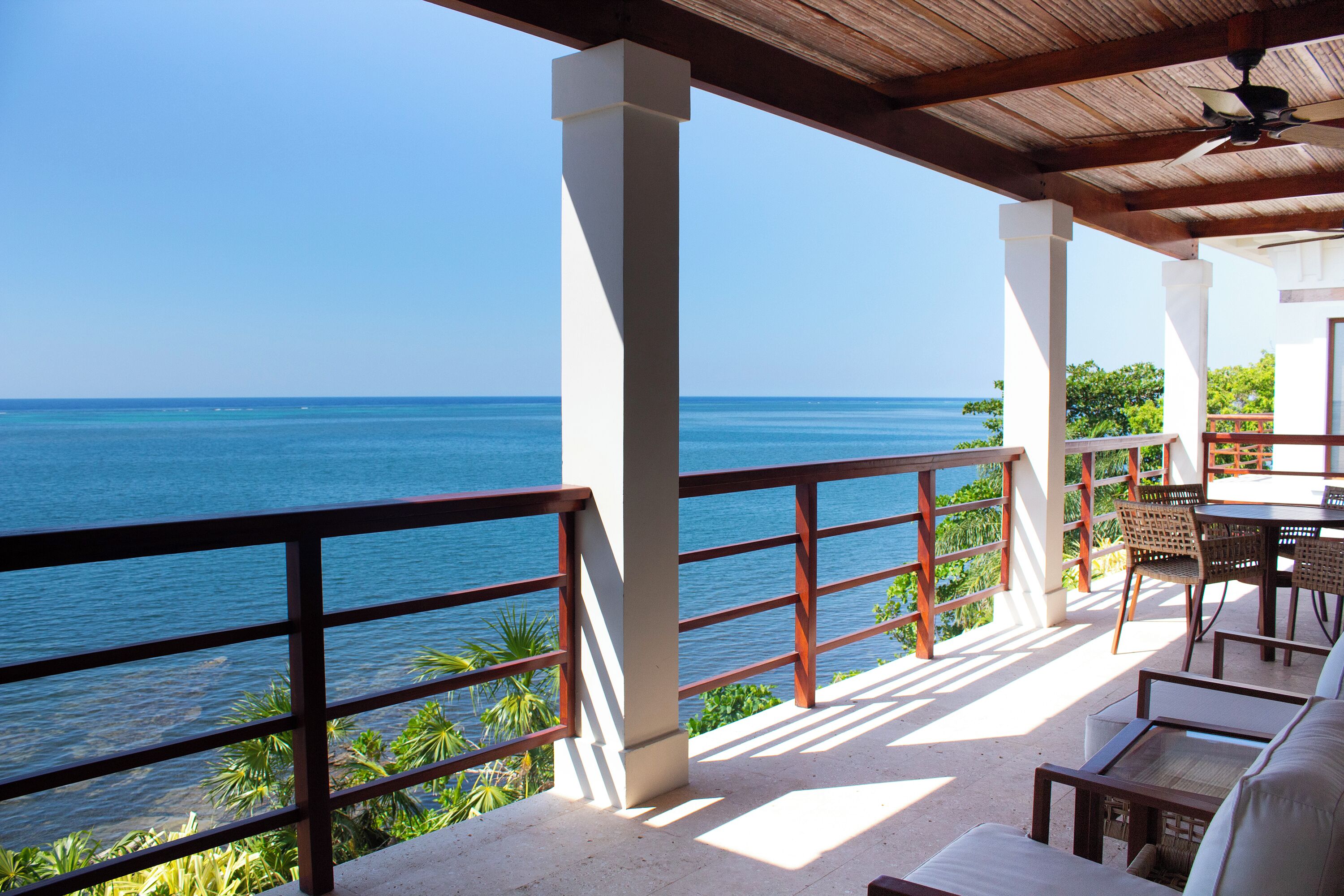 A balcony with a view of the ocean, wooden furniture, and a ceiling fan.