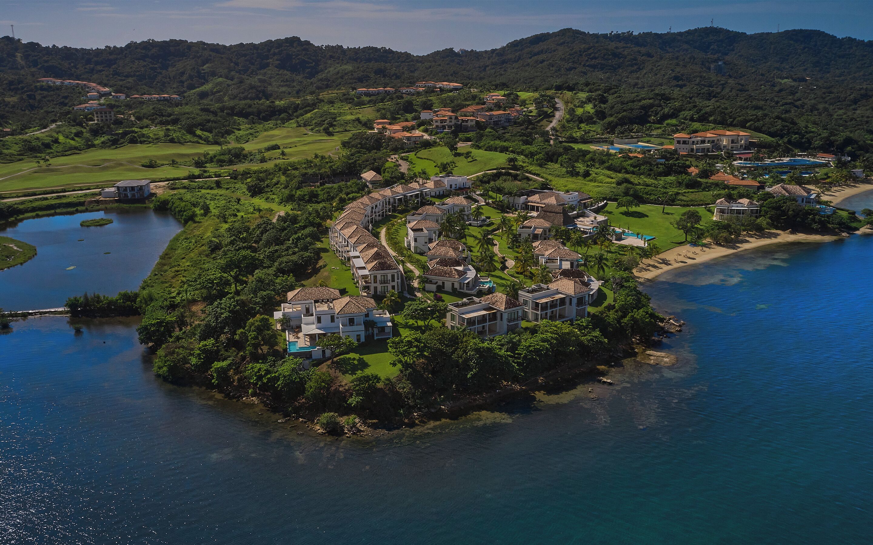 An aerial view of a coastal resort with villas arranged around a central water body, surrounded by lush greenery and a beach.