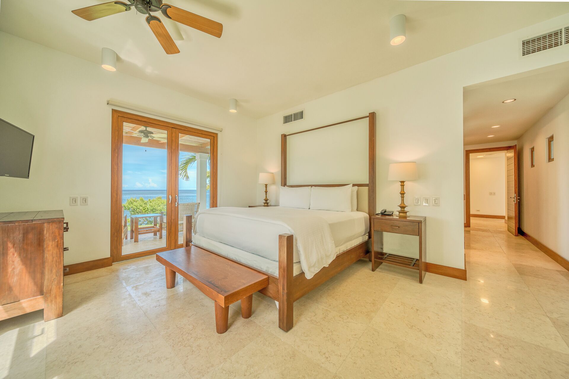 A bedroom with a bed, bedside tables, a bench, a TV, and a view of the ocean.