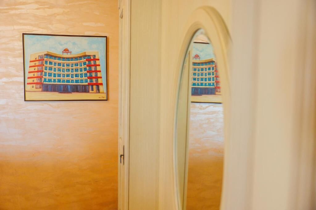 Habitación de hotel con una puerta, un cuadro enmarcado de un edificio en la pared y un espejo que refleja la misma imagen.