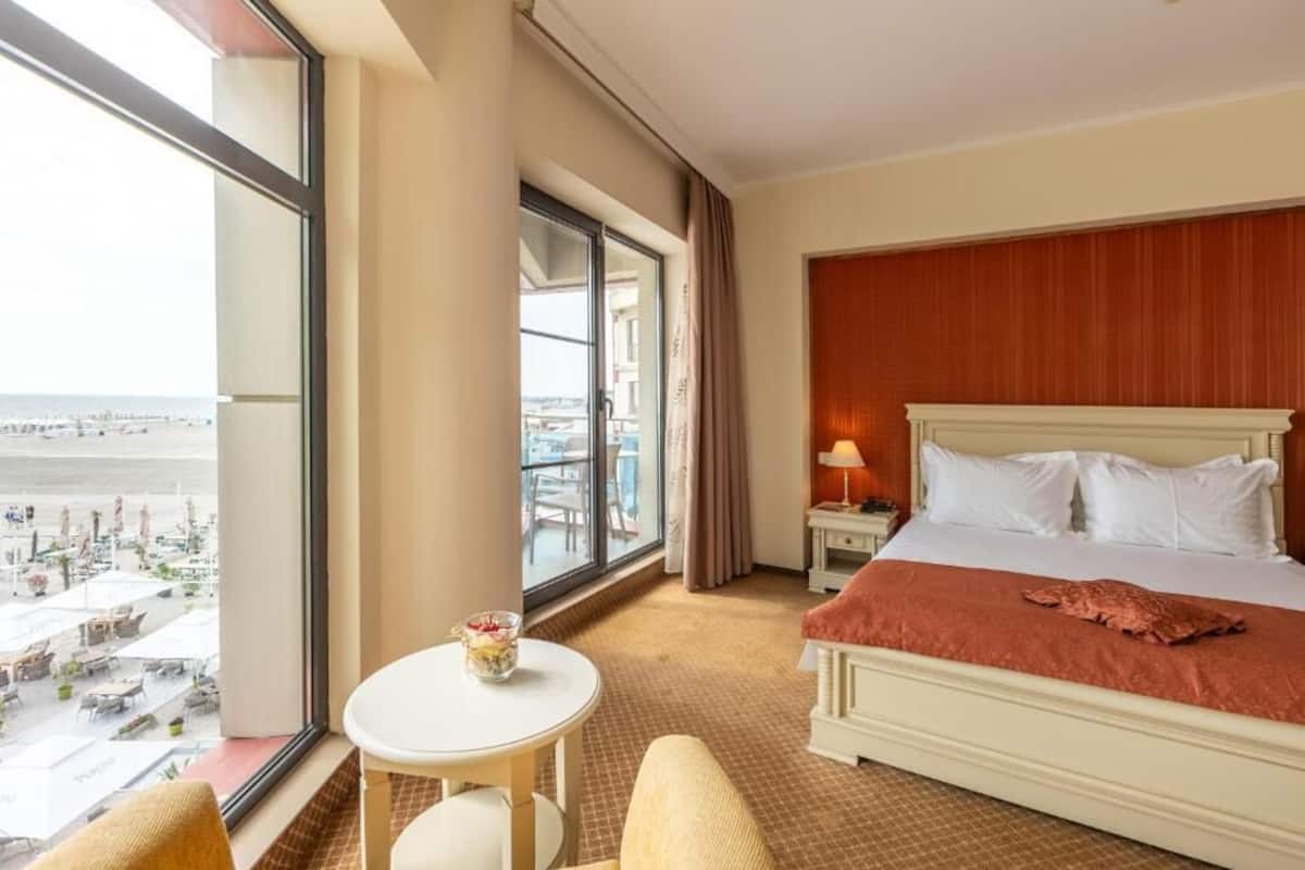Habitación de hotel con una ventana grande, una cama, una mesita con una bebida y vista a la playa.