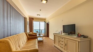 Ein Hotelzimmer mit einer Couch, einem Fernseher auf einer Kommode, einem Esstisch und Meerblick durch große Fenster.