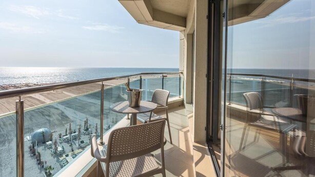 Ein Balkon mit Glasgeländer, zwei Stühle, ein kleiner Tisch und Blick auf das Meer und entfernte Gebäude.
