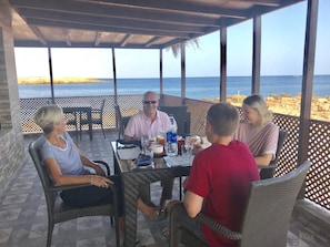 A covered outdoor dining area with a view of the sea, featuring a table with chairs and a few people seated.