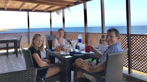 A covered outdoor seating area with wicker furniture, a table with drinks and a bottle, and a view of the ocean.
