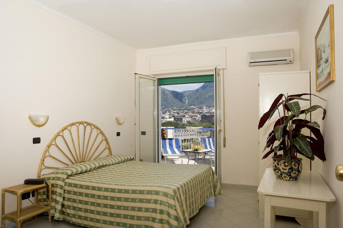 Un dormitorio con cama, una mesita, una planta en maceta y un balcón con vistas a un pueblo y montañas.