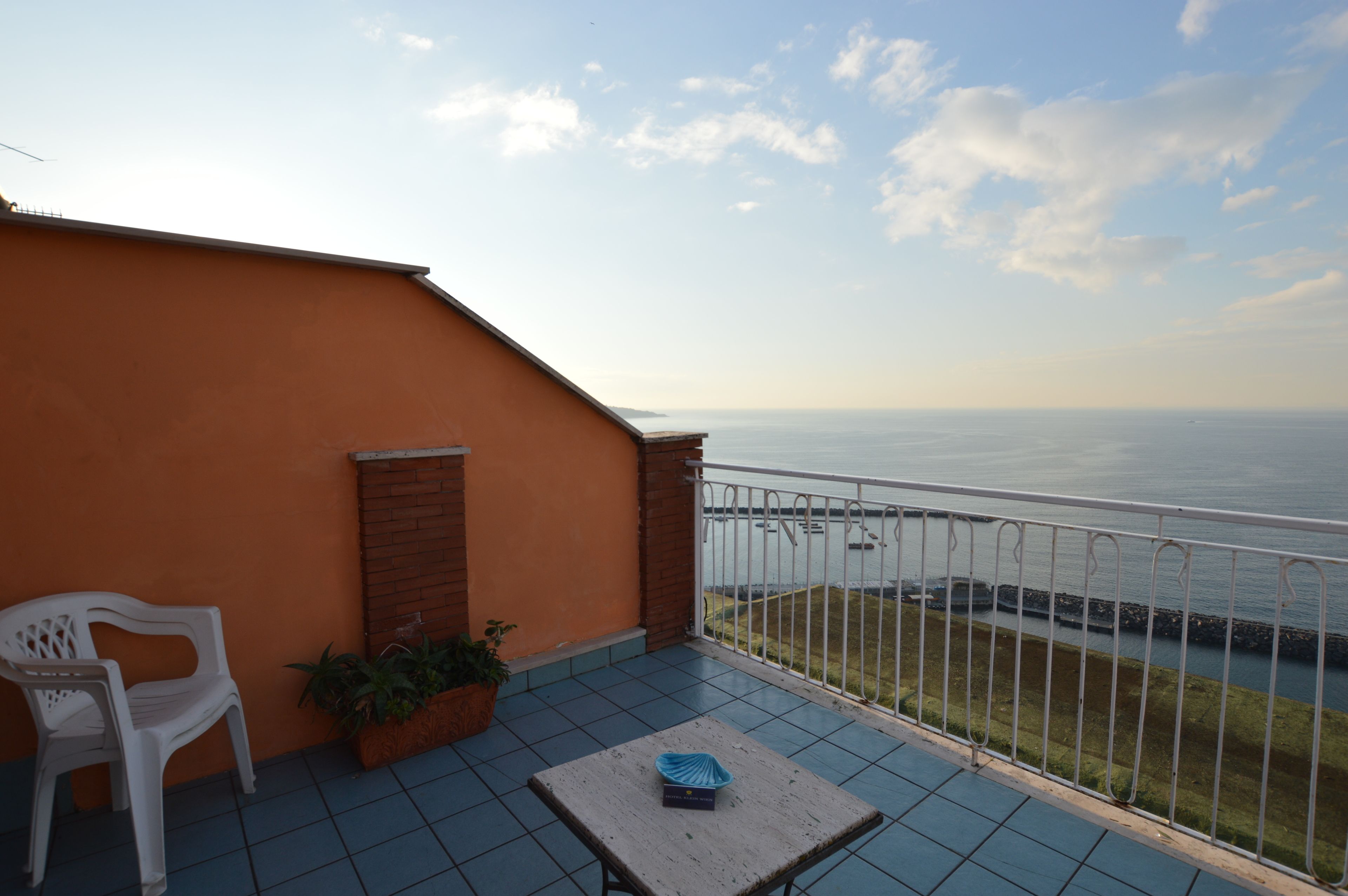 A balcony with a view of the sea, a white plastic chair, a small table, and potted plants.
