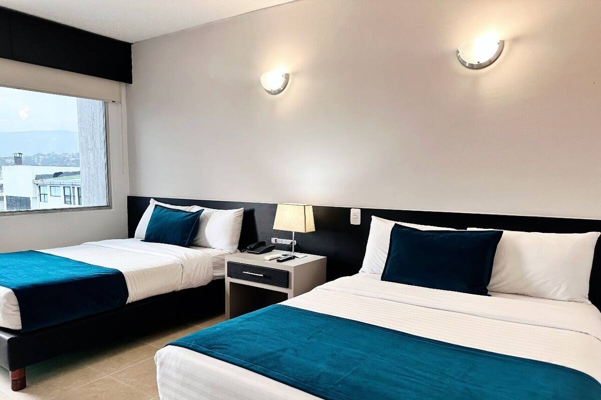 Hotelkamer met twee bedden, elk met een blauwe spreideken en witte lakens. Er is een nachtkastje met een lamp en een telefoon, en een raam met uitzicht op gebouwen.