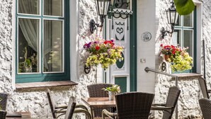 Un edificio con fachada blanca y marcos de ventana verdes, que cuenta con asientos exteriores con sillas de mimbre y una mesa.