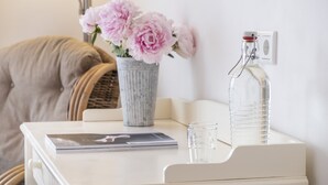 Una mesita de noche blanca con un jarrón de flores rosas, una botella de vidrio transparente y un libro.