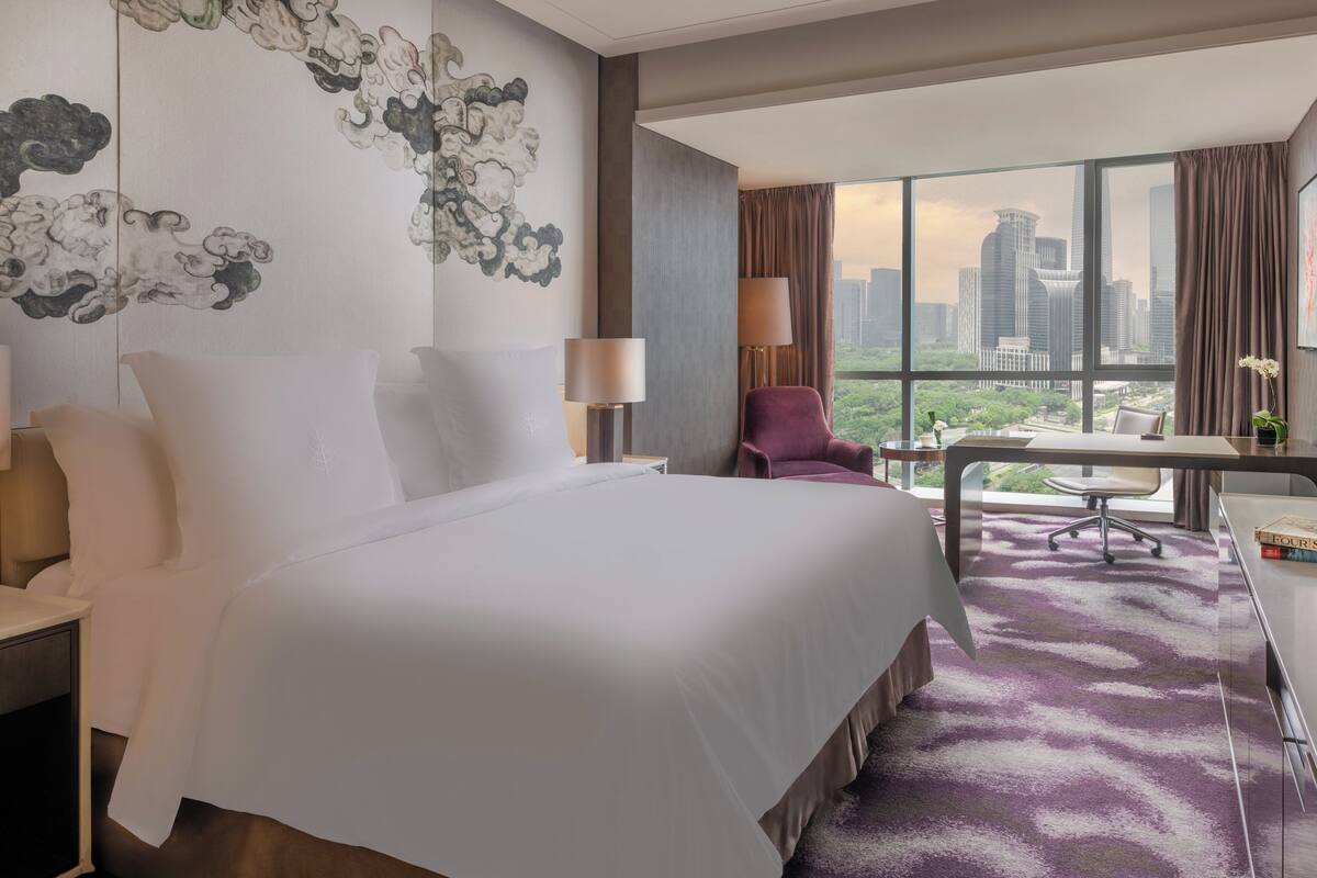 A hotel room with a large bed, a desk, a chair, and a view of a cityscape through large windows.