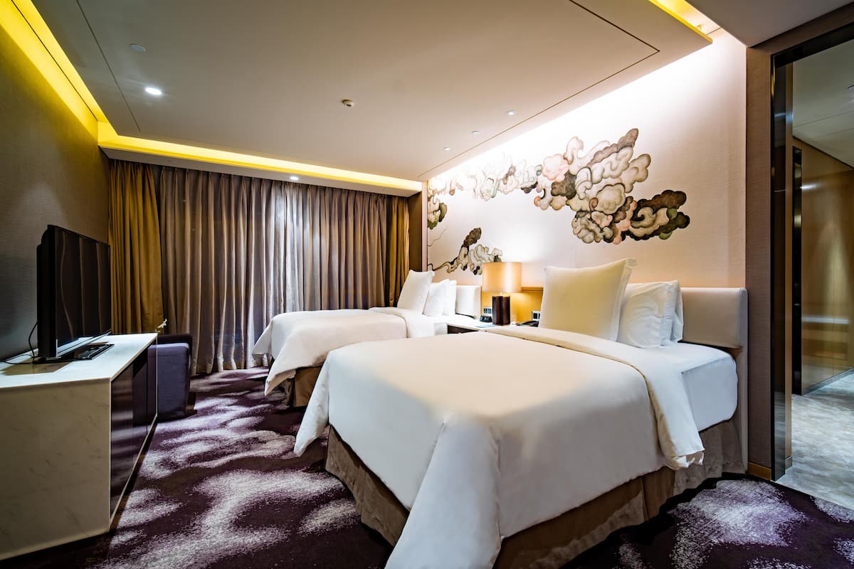 A hotel room with two beds, a desk, a TV, and a large window with curtains.