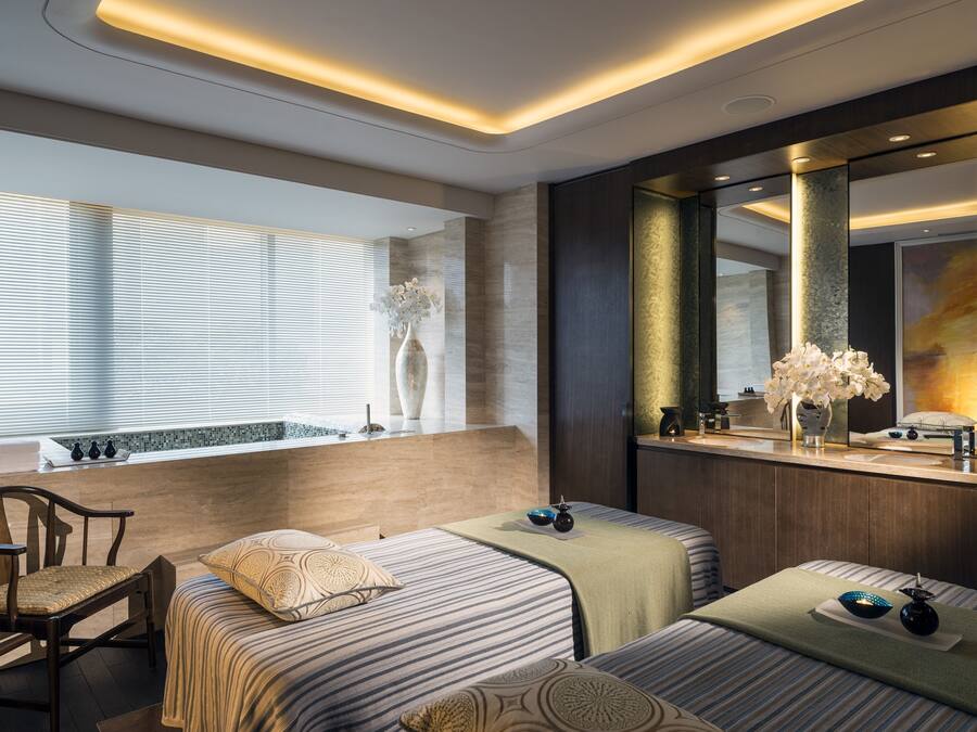 A modern hotel room with a large mirror, two beds with striped bedding, a wooden chair, and a vase of flowers on the windowsill.