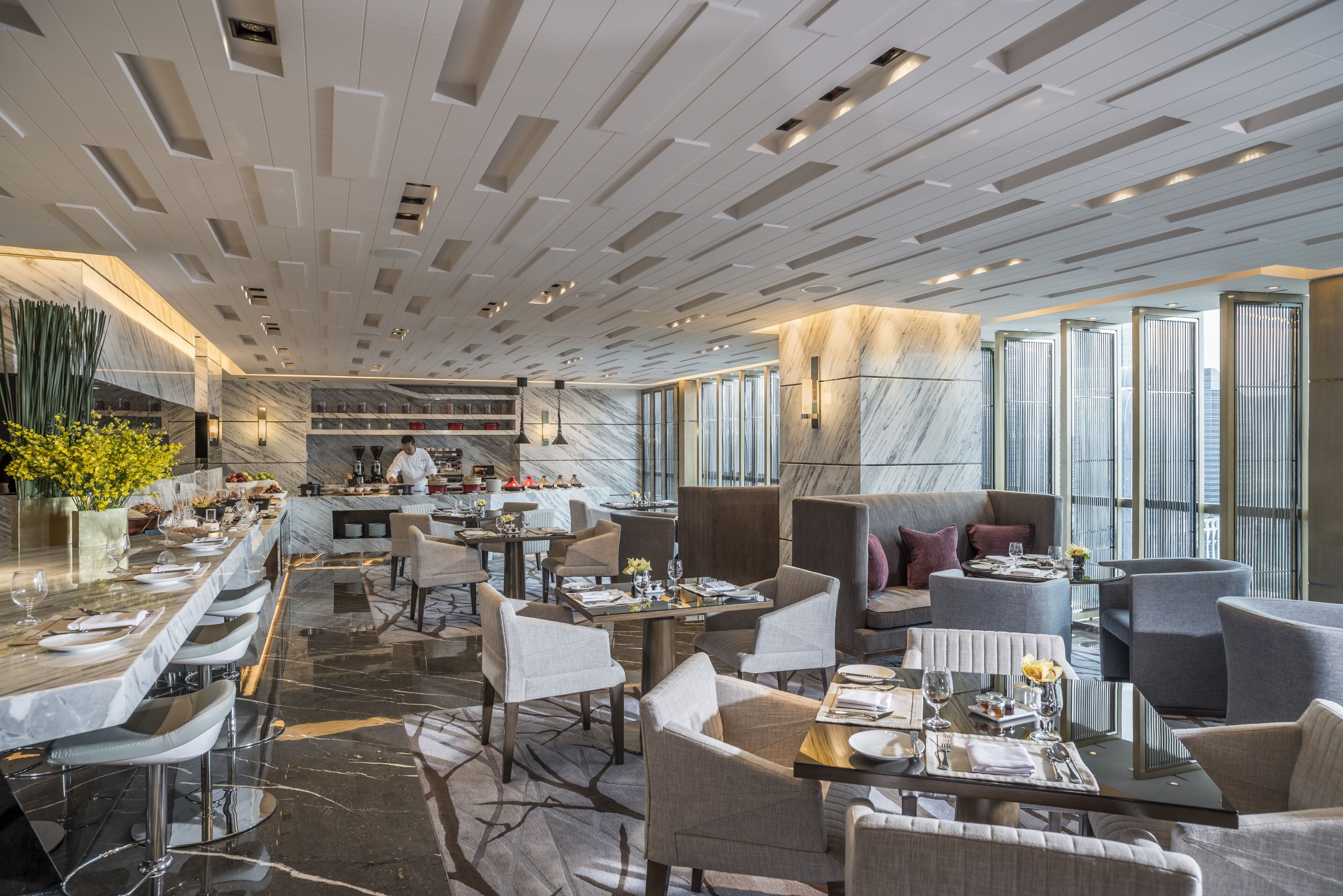 A modern restaurant with a long bar, multiple dining tables, and a chef preparing food.