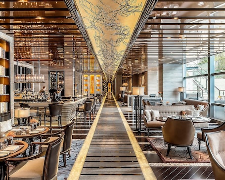 A long, narrow hotel lobby with a central walkway, seating areas on both sides, and a bar on the left.
