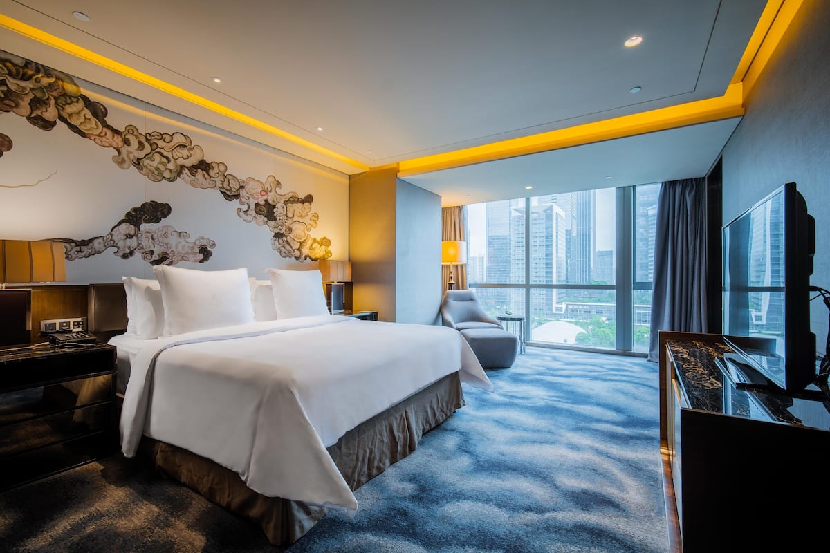 A modern hotel room with a large bed, a desk, a chair, a TV, and a view of the city.