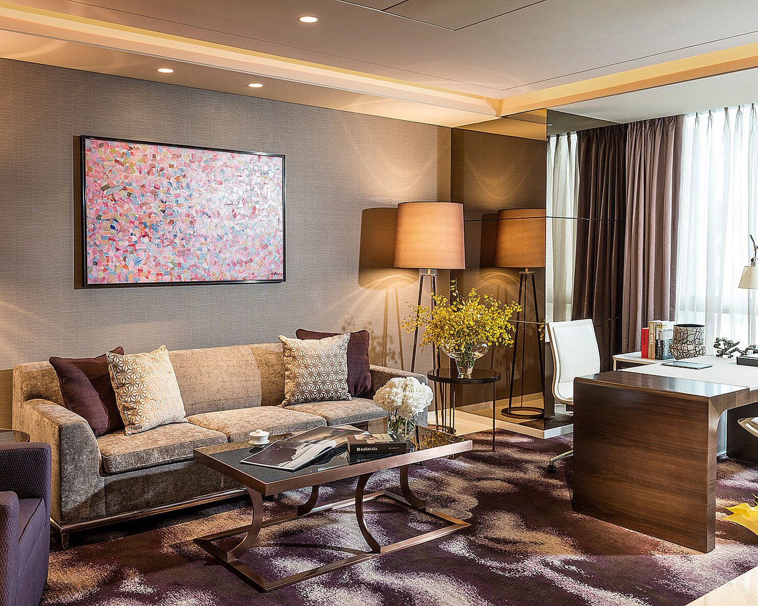 A modern hotel room with a sofa, coffee table, and a desk with a lamp. There's a large abstract painting on the wall and a window with curtains.