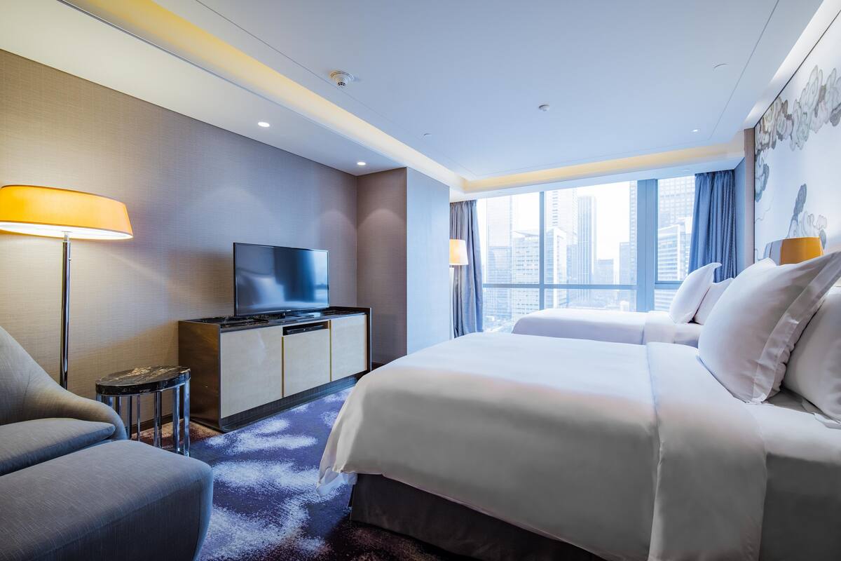A modern hotel room with a large bed, a TV, a desk, and a view of the city.