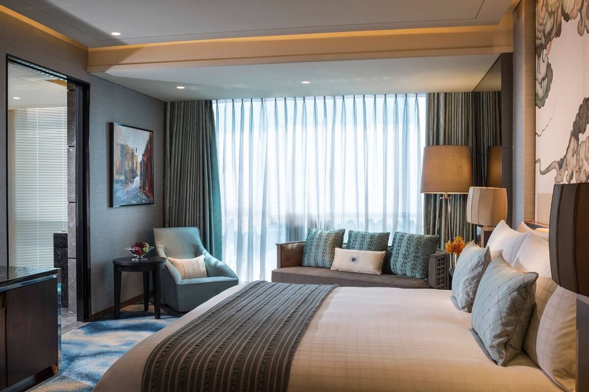 A modern hotel room with a large bed, a TV, a seating area with a sofa and chairs, and a large window with curtains.