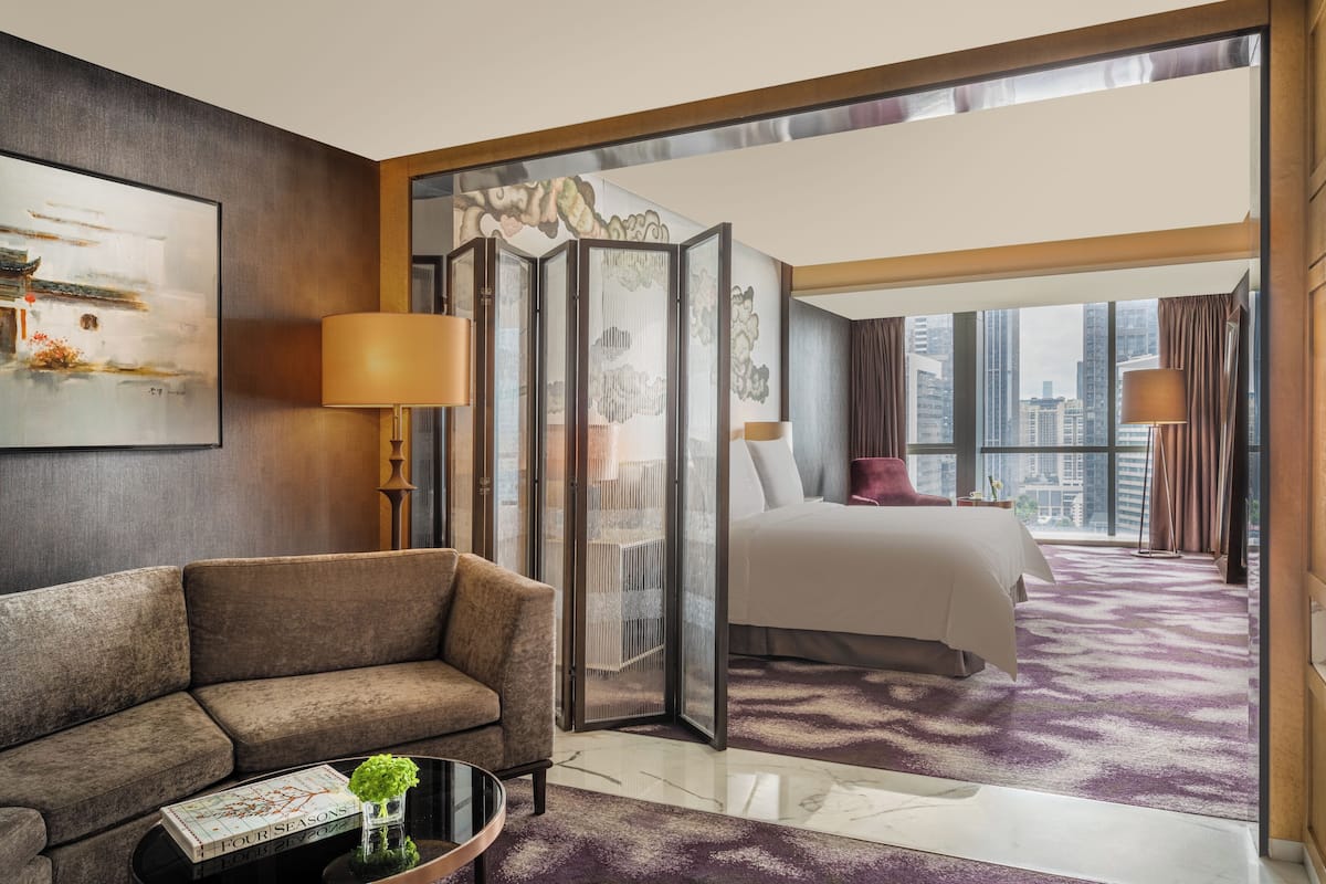 A hotel room with a sofa, a bed, a large window with a city view, a decorative screen, and a lamp.