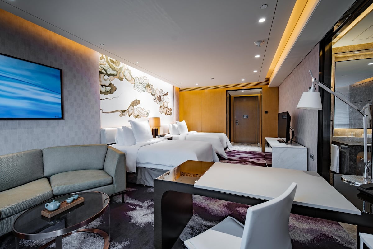 A modern hotel room with a sofa, two beds, a dining table, and a TV.