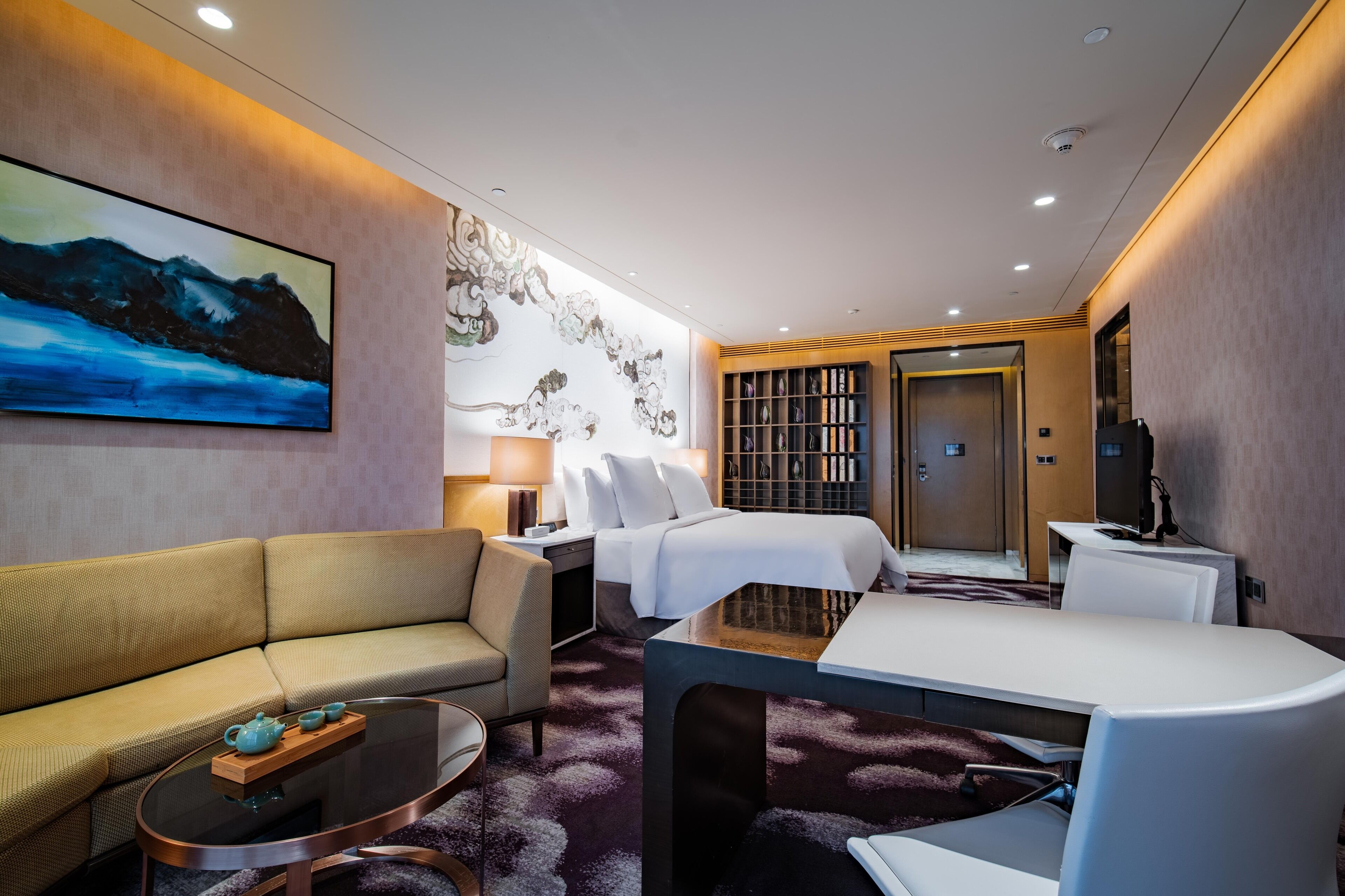 A hotel room with a sofa, a bed, a desk, a bookshelf, and a painting on the wall.
