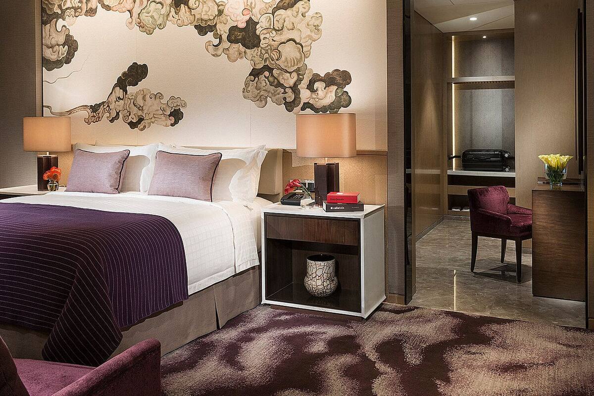 A modern hotel room with a large abstract wall mural, a neatly made bed with multiple pillows, a bedside table with a lamp, a small vase with flowers, and a chair in the corner.